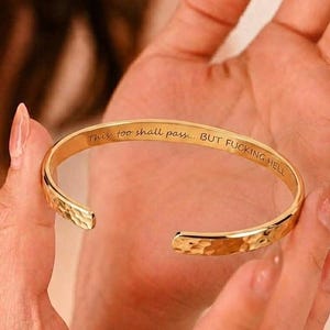 May include: A gold-toned metal cuff bracelet with a hammered exterior texture. The inner surface of the bracelet features the engraved text: This too shall pass... BUT FUCKING HELL. The bracelet has an open design with rounded ends.
