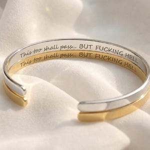 May include: Two open cuff bracelets, one silver-toned and one gold-toned, each engraved with the black text: This too shall pass... BUT FUCKING HELL. The bracelets feature a smooth, polished finish and a simple, minimalist open-ended design.