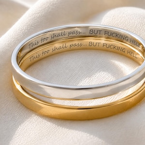 Puede incluir: Dos anillos finos de metal, uno de tono plateado y otro de tono dorado, apilados juntos. La superficie interior de cada anillo está grabada con el texto en negro: This too shall pass... BUT FUCKING HELL. Los anillos tienen un acabado pulido.