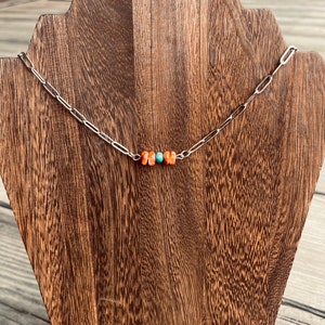 May include: A silver-toned chain necklace with a row of orange and turquoise beads. The necklace is displayed on a wooden stand with a natural wood grain finish. The beads are small and round, adding a pop of color to the simple design.
