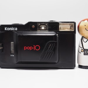 Op de afbeelding: Zwarte Konica pop-10 camera met rode tekst en bijpassende riem. Een houten pop met een camera om de nek staat naast de camera. De camera heeft een flitser en een afstandsinstelling van 1,5-2 m.