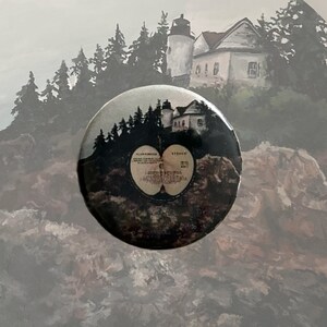 May include: A round button featuring a black and white image of a lighthouse on a rocky cliff with trees and a record album cover in the foreground. The album cover is a half-eaten apple with the text "The Beatles" and "Sgt. Pepper's Lonely Hearts Club Band" printed on it.
