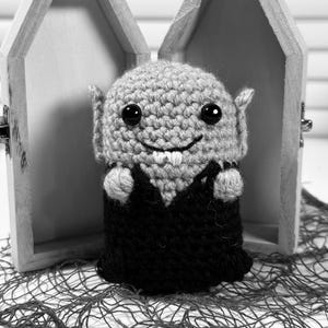 May include: A grayscale image of a crocheted vampire doll with large black eyes, small pointed ears, and a smiling mouth with two small fangs. The doll is wearing a black cape and is positioned in front of an open wooden coffin.