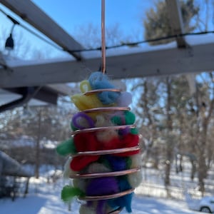 May include: A copper wire bird feeder with a hook, filled with colorful wool yarn. The feeder is hanging from a wooden structure against a snowy background.