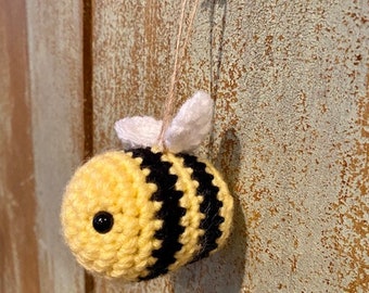 May include: A crocheted bee ornament with black and yellow stripes and white wings. The bee is hanging from a string attached to a nail.