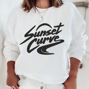May include: White sweatshirt featuring the text "Sunset Curve" in a black, stylised font. The sweatshirt has a crew neck and long sleeves. The text is in a cursive style with a wave design underneath.