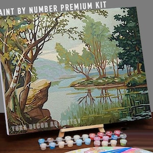 Puede incluir: Kit de pintura por números que presenta una escena de paisaje con un lago, árboles y una costa rocosa. El kit incluye un lienzo, pinturas y pinceles. El lienzo está en un caballete y las pinturas están en pequeños frascos.