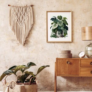 Op de afbeelding: Een botanische print in een gouden lijst toont weelderige groene planten in een decoratieve pot, opgehangen aan een getextureerde muur. Een macram&eacute; wandkleed en een houten bijzettafel met een lamp en klok maken de inrichting compleet. Een grote potplant in een geweven mand staat op de vloer.