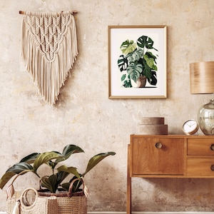 Peut inclure: Int&eacute;rieur de pi&egrave;ce avec une tenture murale en macram&eacute;, une impression botanique encadr&eacute;e et un meuble en bois avec une lampe et une horloge. Une plante en pot dans un panier tress&eacute; est au sol. La palette de couleurs comprend le beige, le marron et le vert.