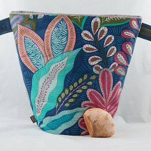 Può includere: Una borsa di tela a forma di tote con motivo floreale blu e verde con un panino marrone all'interno. La borsa ha due manici in tessuto.