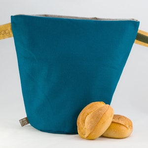 Può includere: Un sacchetto per il pane in tessuto color teal con manici in tessuto giallo e verde. Il sacchetto è etichettato "bunbag" e "GOOD STUFF DTN". Due panini bianchi sono davanti al sacchetto.