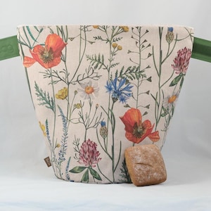 Puede incluir: Una bolsa de tela beige con un estampado botánico de amapolas rojas, acianos azules y tréboles rosas. La bolsa tiene pestañas laterales verdes con cierres de velcro y la palabra burbag grabada. Un pequeño panecillo marrón se sitúa delante.