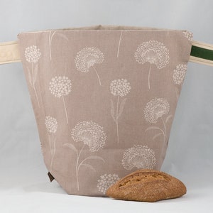 Puede incluir: Una bolsa de pan de lino beige con estampado floral blanco y detalles en cuero marrón. La bolsa tiene dos asas, una con una correa de tela beige y la otra con una correa de tela verde. Un pan está delante de la bolsa.