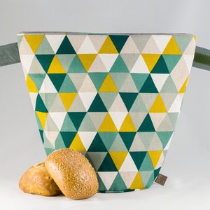 Könnte beinhalten: Eine geometrische Brottasche mit einem Dreiecksmuster in Türkis, Gelb und Weiß. Die Tasche hat graue Griffe mit Klettverschlüssen. Zwei runde Brötchen mit Sesamkörnern befinden sich vor der Tasche.
