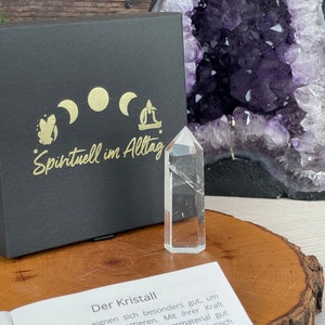 Può includere: Un punto di cristallo di quarzo trasparente su una superficie di legno con una scatola nera e un libro con il testo "Der Kristall" e "Spintuell im Alltag".
