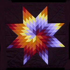 Puede incluir: Un edredón colorido con un diseño de estrella de ocho puntas. Las puntas de la estrella muestran una transición de colores del rojo al naranja, amarillo y morado. El edredón está sobre un fondo oscuro, resaltando los tonos vibrantes de la estrella. El edredón es probablemente hecho a mano.