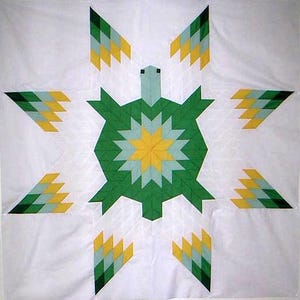 Może przedstawiać: Ręcznie robiona kołdra z centralnym motywem żółwia. Żółw jest zielony z żółtym i jasnoniebieskim środkiem w kształcie gwiazdy. Osiem stylizowanych kształtów strzałek w kolorze zielonym, żółtym i jasnoniebieskim otacza żółwia na białym tle.