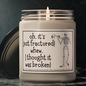 May include: A white soy candle with a black and white illustration of a skeleton with the text "oh. it's just fractured! whew. I thought it was broken!" and "8oz. Soy Candle | 50-60 Hour Burn Time | Design by Kelley Ann Co."