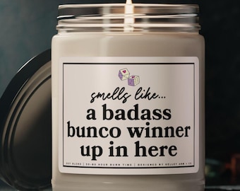 Puede incluir: Una vela blanca con una tapa negra. La etiqueta de la vela dice "smells like... a badass bunco winner up in here".