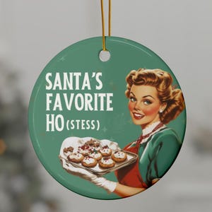 Könnte beinhalten: Ein runder, grüner Weihnachtsschmuck mit dem Text "SANTA'S FAVORITE HO (STESS)". Das Ornament zeigt eine Vintage-Illustration einer Frau, die ein Tablett mit Keksen hält. Der Anhänger hat eine goldene Schnur zum Aufhängen.