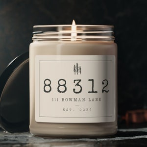 May include: A beige candle in a glass jar with a white label. The label has a black silhouette of trees and the text "88312 111 BOWMAN LANE EST. 2024".