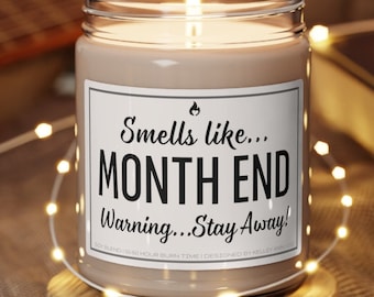 May include: A lit candle in a glass jar with a label that reads "Smells like... MONTH END Warning... Stay Away!". The candle sits on a wooden coaster, surrounded by string lights.