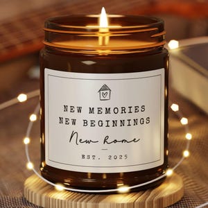 May include: A lit brown glass jar candle with a white label that reads "NEW MEMORIES NEW BEGINNINGS New Home EST. 2025". The candle sits on a wooden coaster, surrounded by string lights.