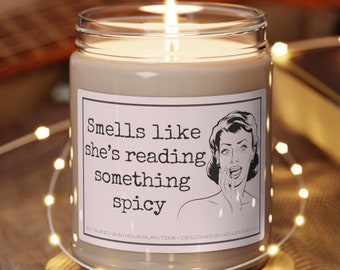 May include: A soy candle in a clear glass jar with a white label that reads "Smells like she's reading something spicy". The label also includes a retro-style illustration of a woman with a surprised expression. The candle is sitting on a wooden coaster.