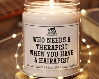 May include: A white candle with a glass jar and a black label that reads "WHO NEEDS A THERAPIST WHEN YOU HAVE A HAIRAPIST". The label also includes the text "8 oz. | 50-60 HOUR BURN TIME | KELLEY ANH.CO".