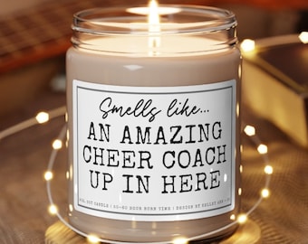 May include: A brown soy candle with a white label that reads "Smells like... AN AMAZING CHEER COACH UP IN HERE". The candle is lit and sitting on a wooden coaster.