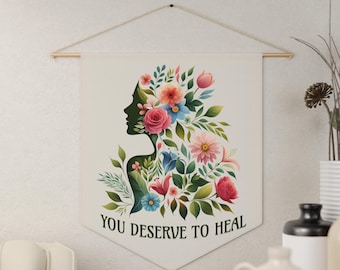 Peut inclure: Une tapisserie murale en toile blanche avec une silhouette de femme avec des fleurs dans les cheveux. Le texte "You Deserve to Heal" est imprimé sous l'image.