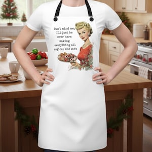 Puede incluir: Delantal blanco con correas negras. Presenta una ilustraci&oacute;n vintage de una mujer sosteniendo un plato de galletas y el texto "Don't mind me, I'll just be over here making everything all magical and shit."