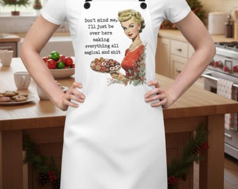 Pode incluir: Avental branco com alças pretas. Apresenta uma ilustração vintage de uma mulher segurando um prato de biscoitos e o texto "Don't mind me, I'll just be over here making everything all magical and shit."