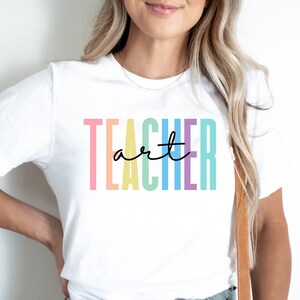May include: White t-shirt with the word "TEACHER" in pastel vertical stripes and the word "art" in a black script font. The shirt is a crew neck and has short sleeves.
