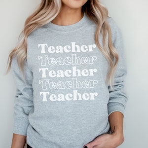 Puede incluir: Una sudadera gris con la palabra "Teacher" repetida cinco veces en una fuente retro blanca.