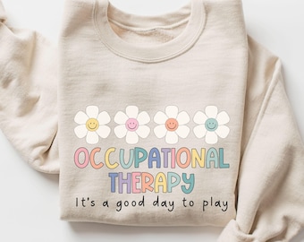 May include: Beige sweatshirt with a crew neck. The front features four white daisy flowers with smiling faces. Below the flowers, the words "OCCUPATIONAL THERAPY" are in colorful letters. The phrase "It's a good day to play" is below.