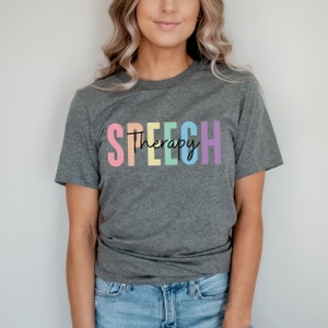 Puede incluir: Una camiseta gris con el texto "SPEECH Therapy" en colores arco&iacute;ris.