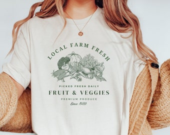 Puede incluir: Camiseta color crema con un gráfico verde de varias frutas y verduras. El texto dice "LOCAL FARM FRESH, FRUIT & VEGGIES, PREMIUM PRODUCE, Since 1989". La camiseta se usa con jeans azules y una rebeca color canela.