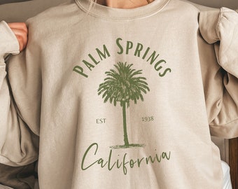Puede incluir: Una sudadera beige con un gráfico de palmera y el texto "Palm Springs Est. 1938 California".