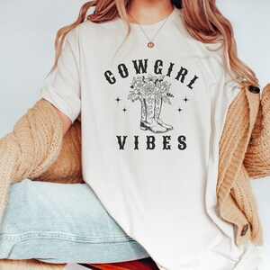 May include: A white t-shirt with a black graphic design of cowboy boots with flowers and the text "COWGIRL VIBES".