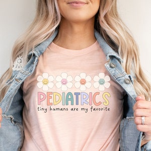 Pu&ograve; includere: Una maglietta rosa con il testo "PEDIATRICS tiny humans are my favorite" in colori arcobaleno. Il testo &egrave; circondato da una fila di cinque margherite bianche con centri gialli e facce sorridenti.