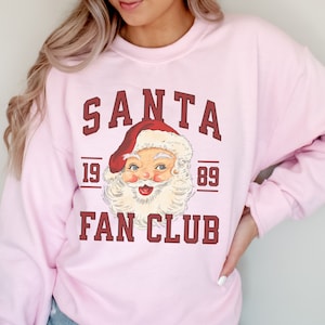 May include: Pink sweatshirt with a vintage Santa Claus graphic and the text "SANTA FAN CLUB 19 89".