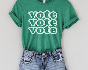 May include: A green t-shirt with the word "vote" repeated three times in a retro font style. The shirt is on a white background and is paired with denim shorts.