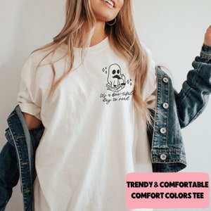 May include: Cream-colored t-shirt featuring a black ghost reading a book, with the text "It's a boo-tiful day to read." The shirt has a relaxed fit and is paired with a denim jacket. The design includes sparkling star accents.