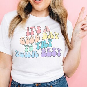 Peut inclure: T-shirt blanc avec un graphique arc-en-ciel qui dit "C'est un bon jour pour essayer de faire de son mieux".