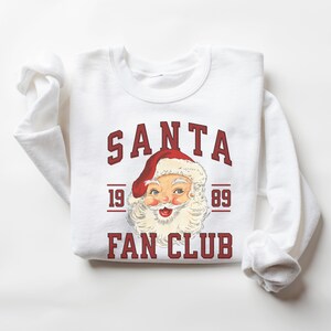 May include: White sweatshirt with a red and white Santa Claus graphic and the text "SANTA FAN CLUB 19 89" in red.