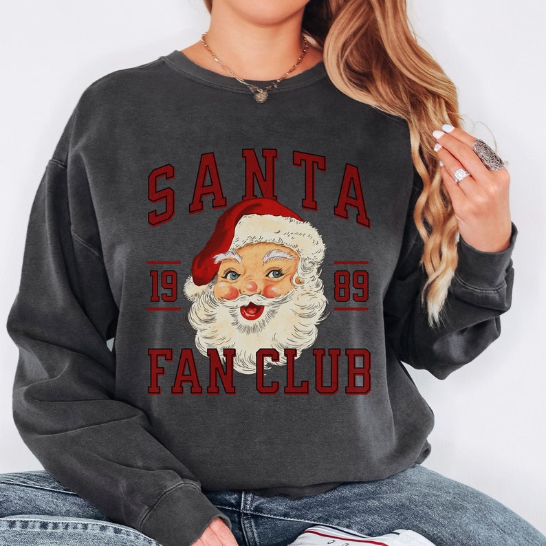 May include: A charcoal gray sweatshirt with a vintage-style graphic of Santa Claus's face. The graphic includes the text "SANTA FAN CLUB" and the numbers "19 89" above and below the image.