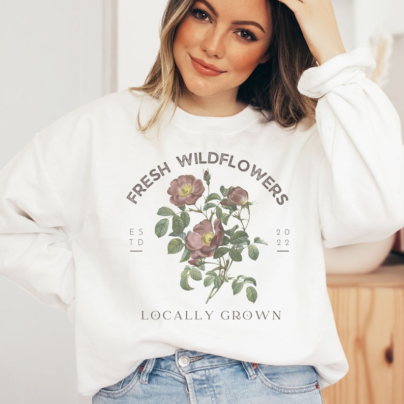 May include: White sweatshirt with a graphic design of a bouquet of pink wildflowers with green leaves. The text "Fresh Wildflowers" is in a circle around the flowers. The text "Est. 2022" is below the flowers. The text "Locally Grown" is below the text "Est. 2022".