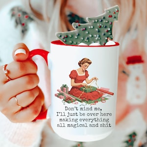 May include: White ceramic mug with a red interior and handle, featuring a vintage illustration of a woman wrapping gifts. The mug has the text "Don't mind me, I'll just be over here making everything all magical and shit." A green Christmas tree cookie sits on the rim.