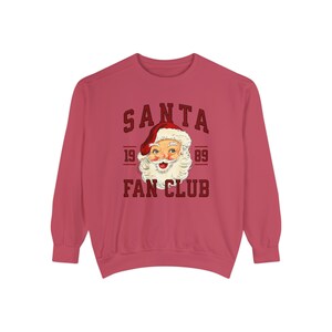 May include: A red long-sleeved sweatshirt with the text "SANTA FAN CLUB" in a red and white vintage style font. The text is surrounded by a red circle with the numbers "19 89" on either side. A cartoon image of Santa Claus's face is inside the circle.